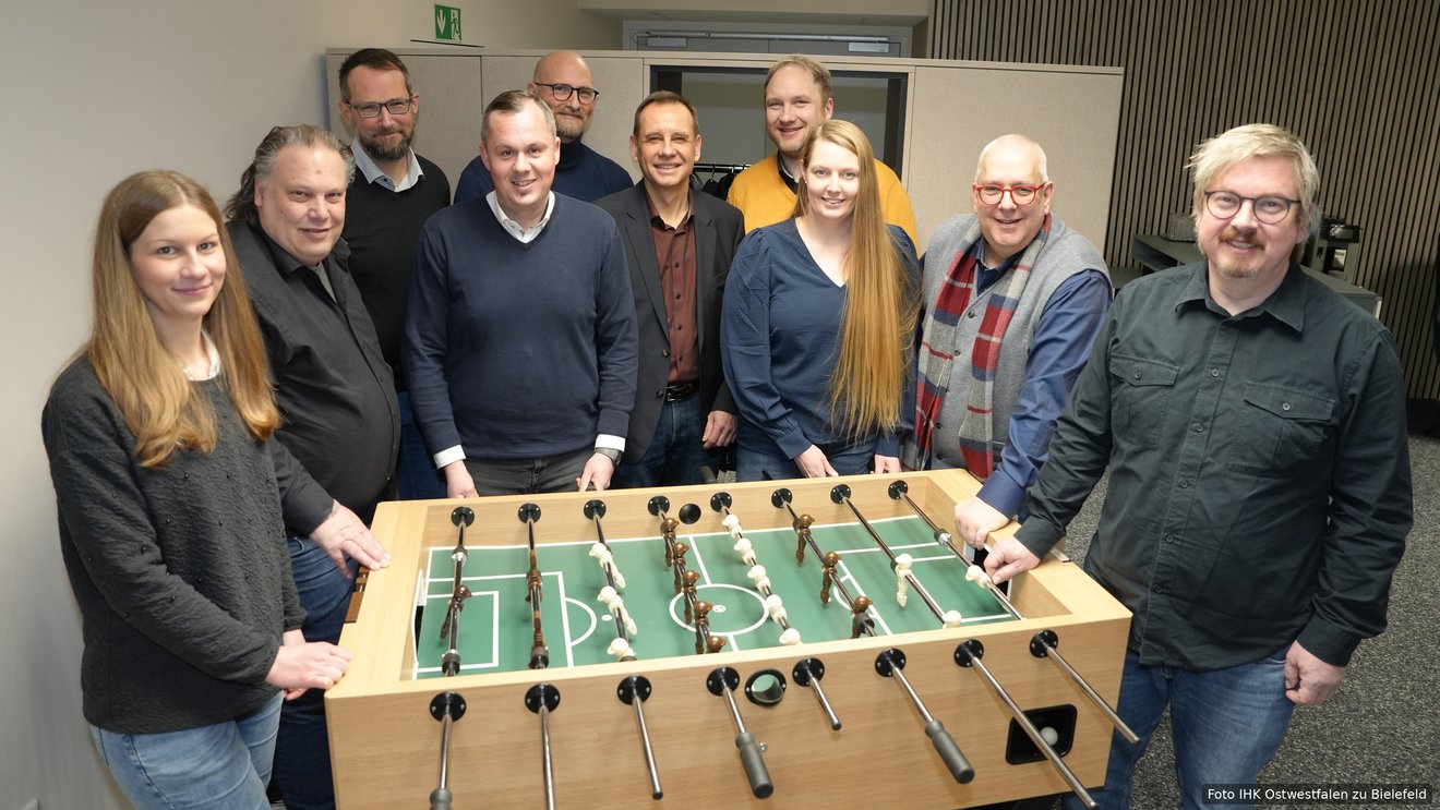 Zehn Personen stehen lächelnd um einen Tischkicker aus hellem Holz in einem Büroraum und blicken in die Kamera.