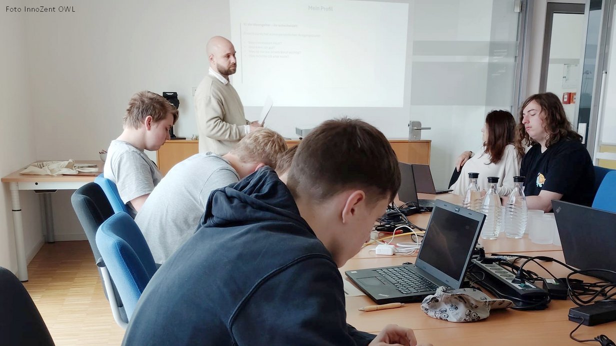 Referent steht vor Präsentationsfolie 'Mein Profil', Jugendliche arbeiten im Vordergrund an Laptops