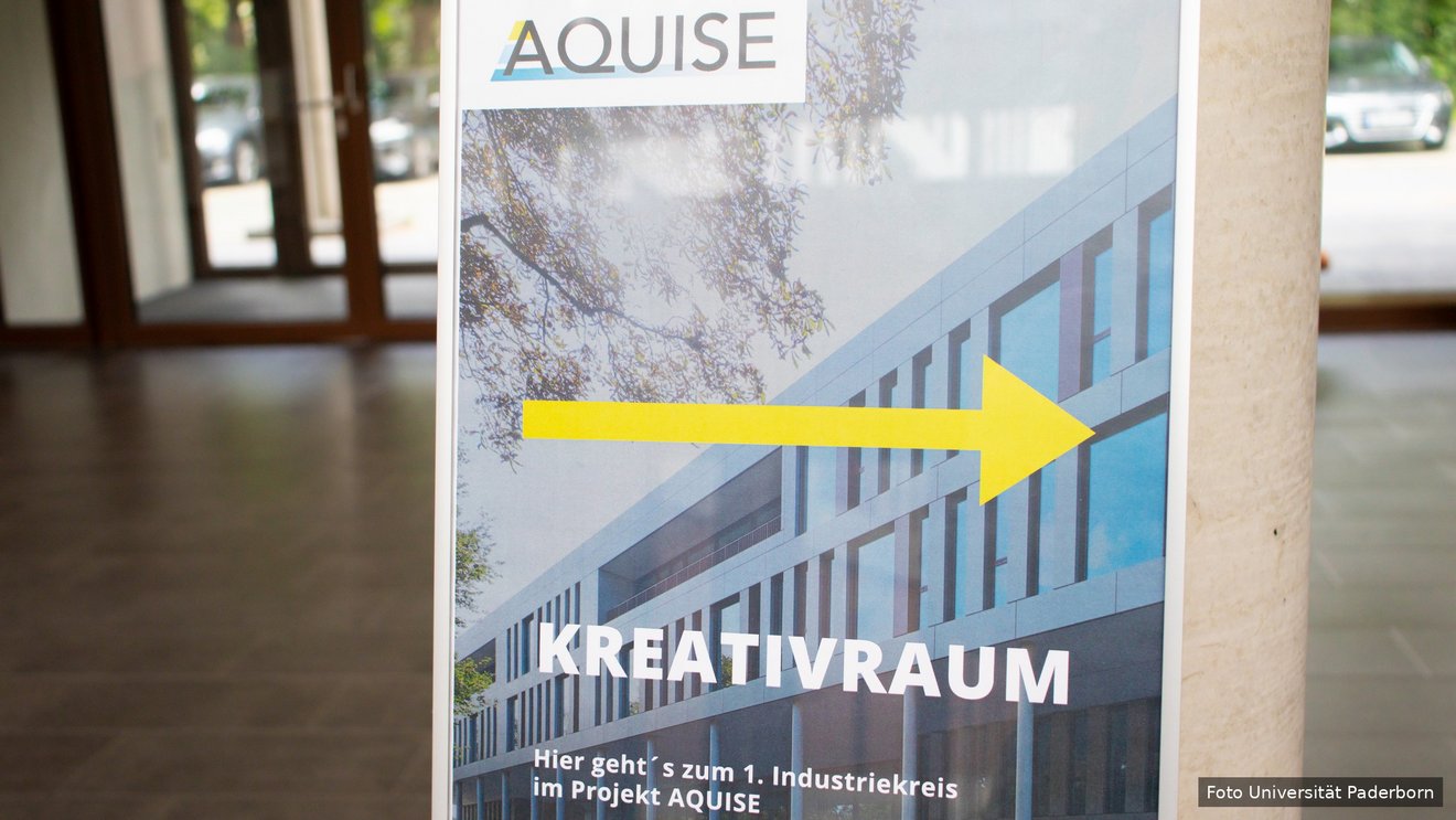 Aufsteller im Foyer mit dem AQUISE-Logo, einem Gebäudefoto und einem gelben Pfeil nach rechts. Darauf steht ‚Kreativraum – Hier geht’s zum 1. Industriekreis im Projekt AQUISE’.