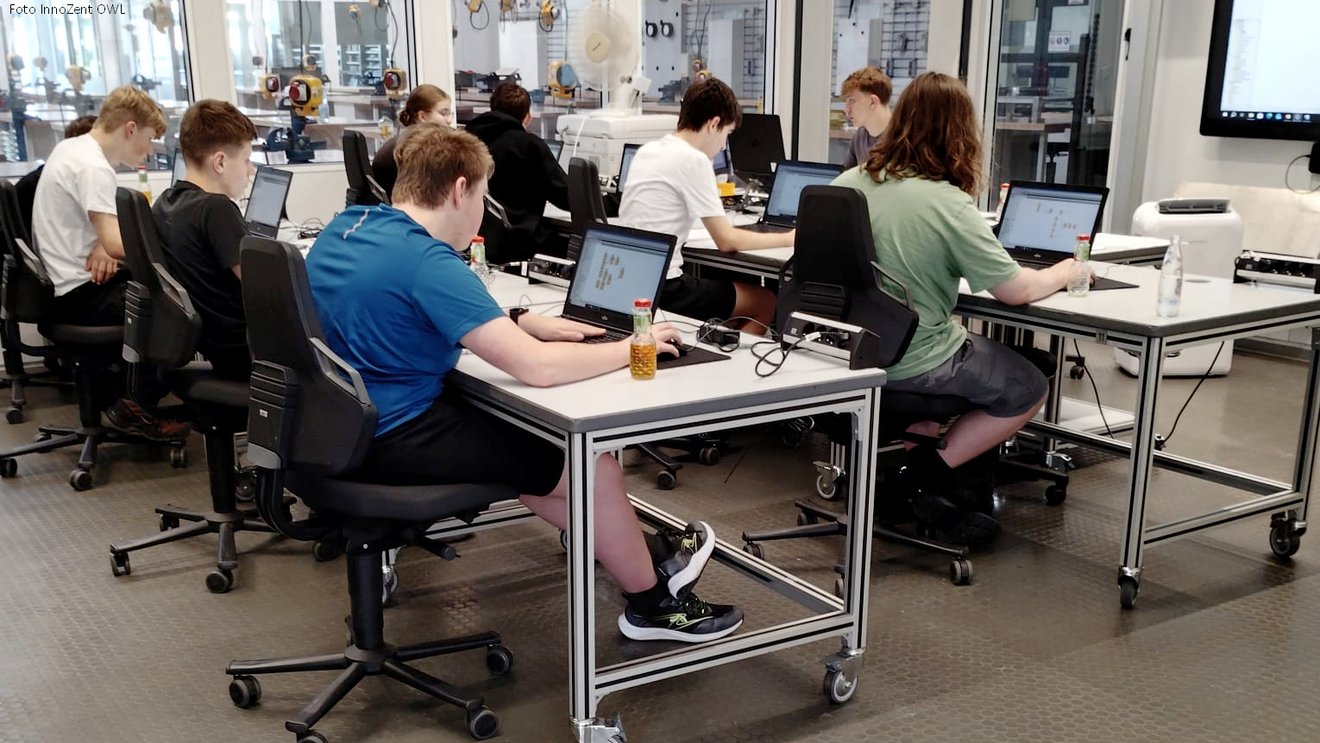 Jugendliche arbeiten konzentriert an Laptops in einem Technik-Workshop mit Robotik- und Elektronikbauteilen im Hintergrund