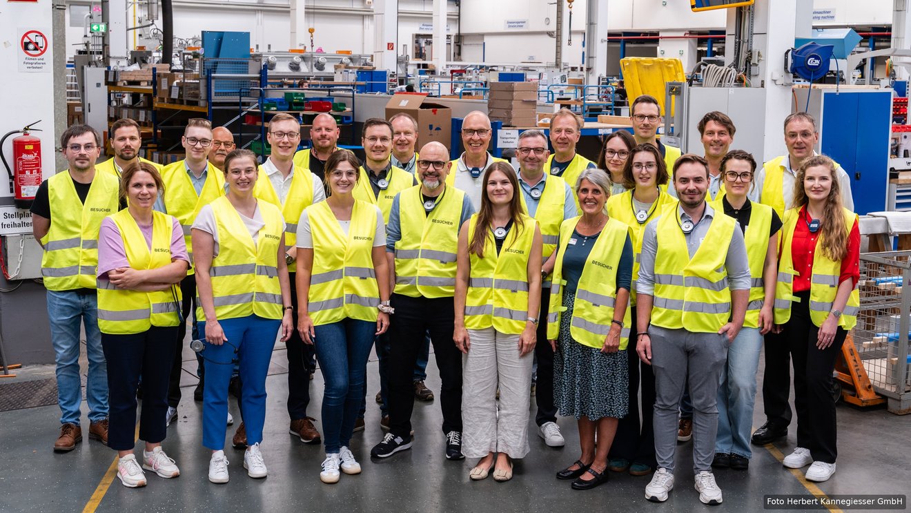 Gruppenfoto: Etwa 25 Personen in gelben Besucherwesten stehen lächelnd in einer Produktionshalle und blicken in die Kamera.