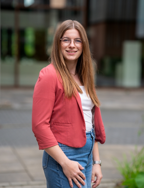 Porträtfoto einer lächelnden jungen Frau mit Brille und langen Haaren, in rostrotem Blazer, die vor einem modernen Bürogebäude steht.