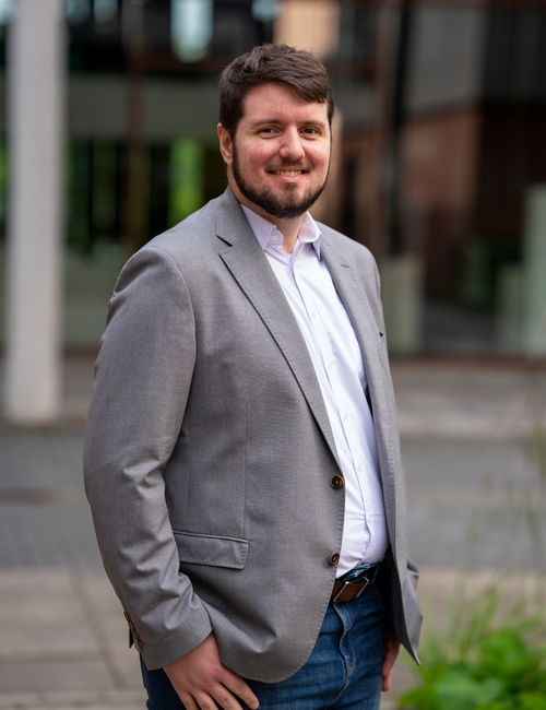 Porträtfoto eines lächelnden Mannes mit Bart, in grauem Sakko und hellem Hemd, der vor einem modernen Bürogebäude steht.