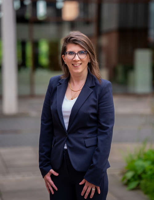 Porträtfoto einer lächelnden Frau mit Brille in dunkelblauem Blazer, die vor einem modernen Bürogebäude steht.