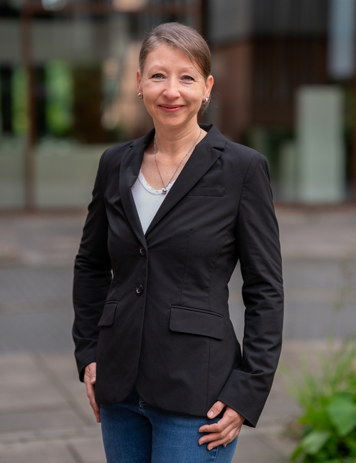 Porträtfoto einer lächelnden Frau in schwarzem Blazer und weißem Top, die vor einem modernen Bürogebäude steht.
