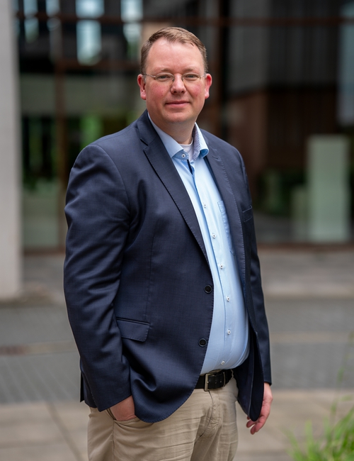 Porträtfoto eines Mannes mit Brille in dunkelblauem Sakko und hellblauem Hemd, der vor einem modernen Bürogebäude steht.