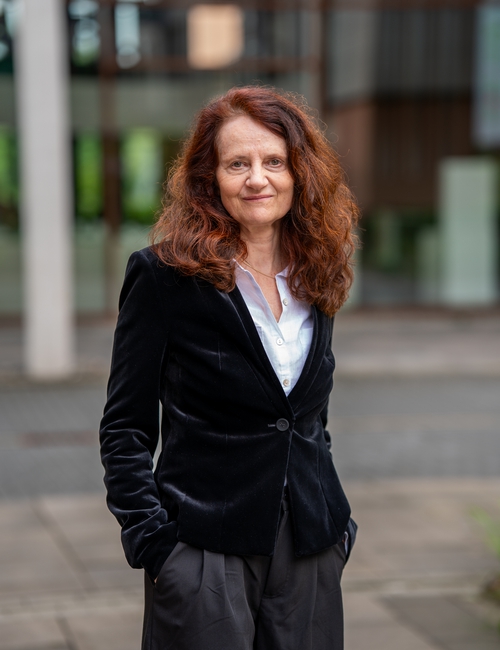 Porträtfoto einer lächelnden Frau mit lockigen rotbraunen Haaren, in schwarzem Samtblazer, die vor einem modernen Bürogebäude steht.