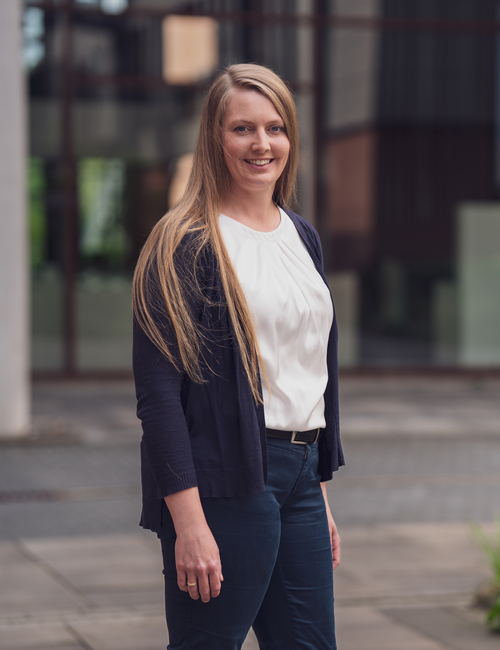 Porträtfoto einer lächelnden Frau mit langen blonden Haaren, in weißer Bluse und dunkler Strickjacke, die vor einem modernen Bürogebäude steht.
