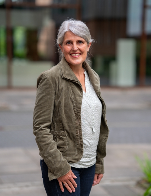 Porträtfoto einer lächelnden Frau mit grauen Haaren, in olivgrüner Jacke, die vor einem modernen Bürogebäude steht.