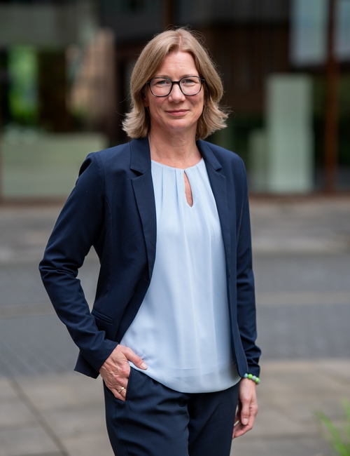 Porträtfoto einer lächelnden Frau mit Brille und schulterlangen blonden Haaren, in dunkelblauem Blazer, die vor einem modernen Bürogebäude steht.