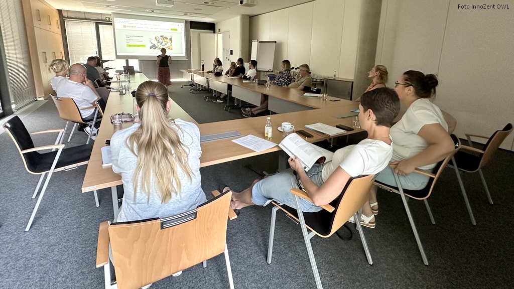 Teilnehmende sitzen an U-förmig angeordneten Tischen und verfolgen einen Vortrag mit Präsentation auf der Leinwand